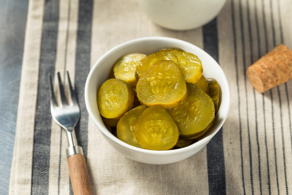 Sliced pickles in a white bowl with a fork on a striped table cloth.