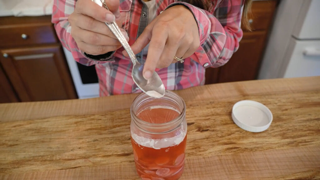 A woman using a spoon to remove kahm yeast from the surface of a ferment in a Mason jar.