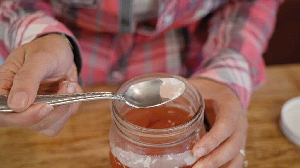 A woman using a spoon to remove kahm yeast from the surface of a ferment in a Mason jar.