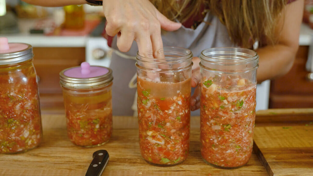 A woman's hand pressing a fermenting weight into a Mason jar of fresh salsa.