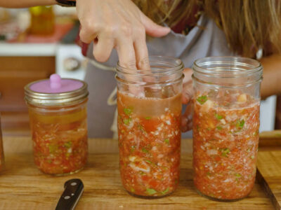 A woman's hand pressing a fermenting weight into a Mason jar of fresh salsa.