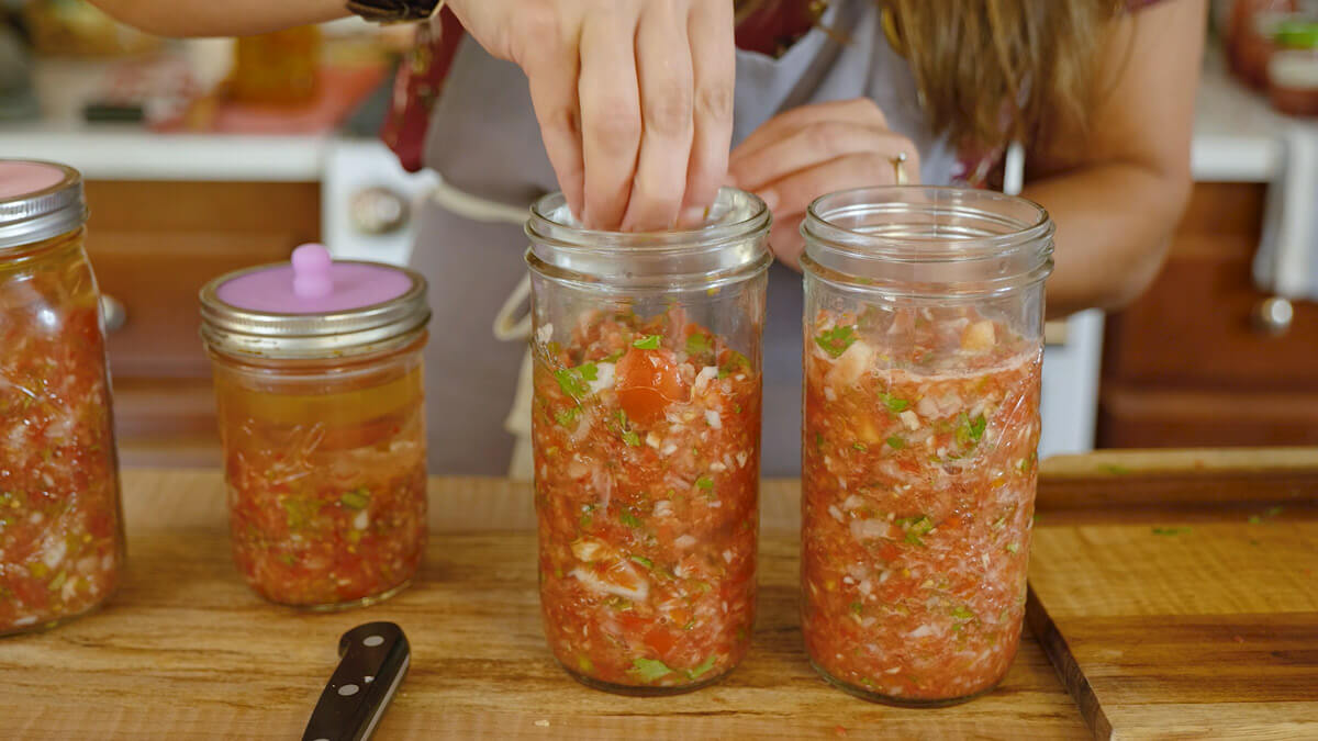 A woman's hand dropping a fermenting weight into a Mason jar of salsa.