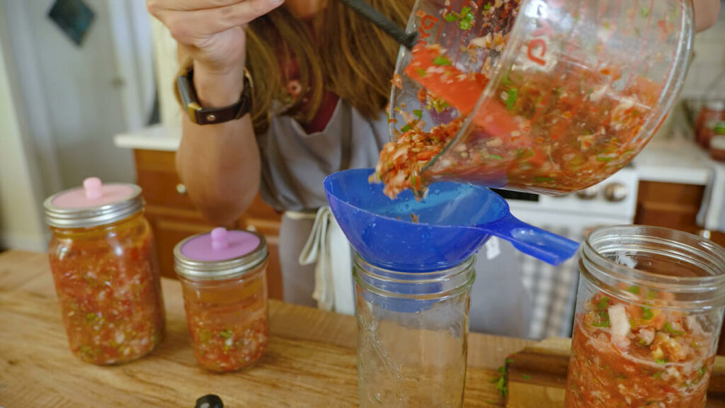 A woman adding fresh salsa to a jar with a blue funnel.