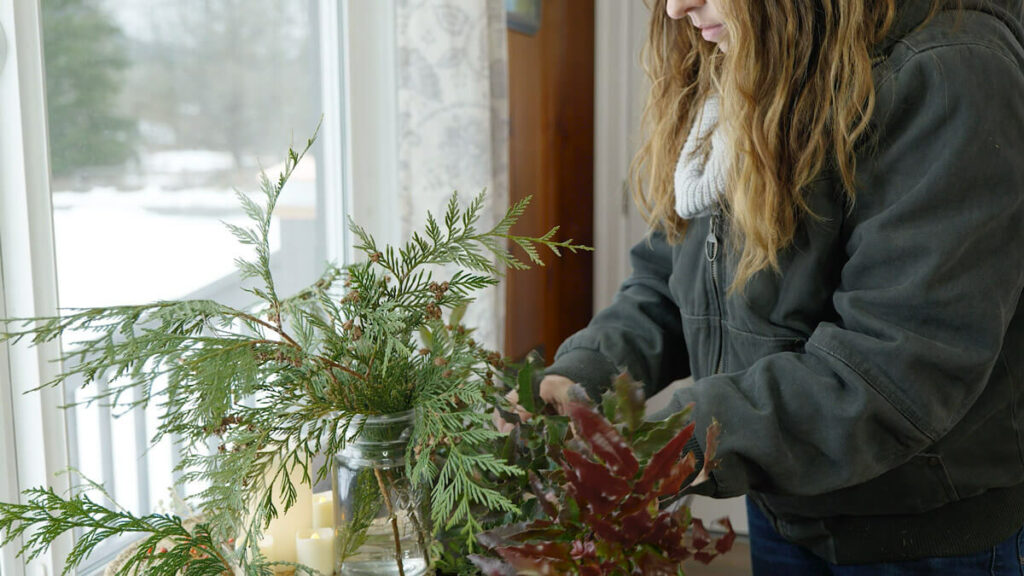A woman arranging an evergreen bouquet in a Mason jar.