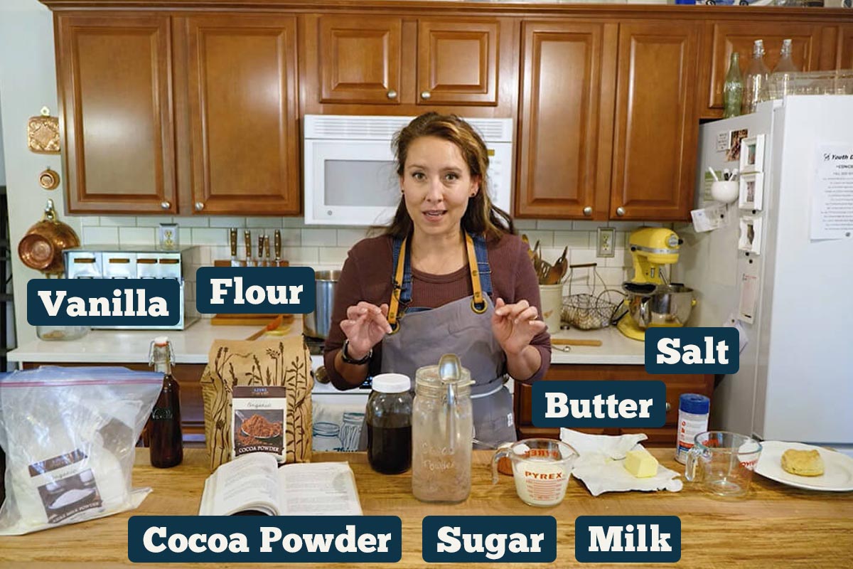 A woman standing in front of chocolate gravy ingredients: flour, butter, salt, cocoa powder, sugar, and milk.