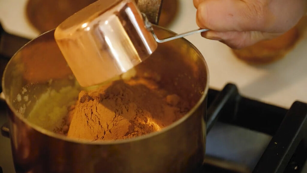 A pot on the stove with cocoa powder being poured in.