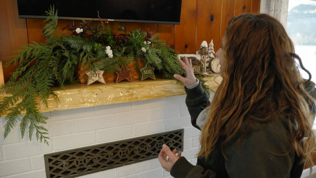 A woman standing by a fireplace mantle with a holiday garland on top.