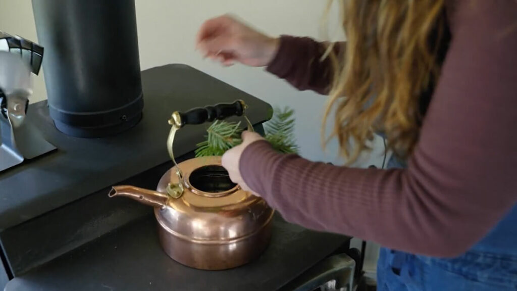 A woman adding evergreen branches to a tea kettle on top of a wood burning stove.