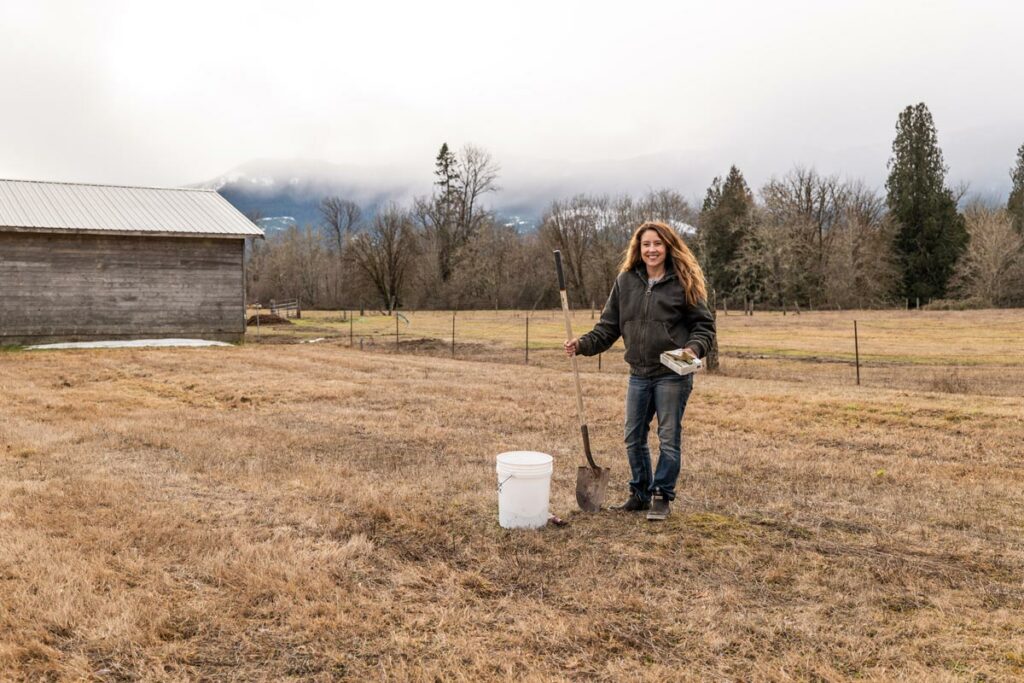 A woman in a field doing a soil test. Holding a shovel next to a bucket.