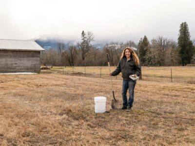 A woman in a field doing a soil test. Holding a shovel next to a bucket.
