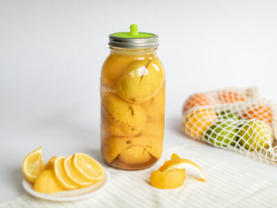 Jar of fermented lemons on a counter with a bag of citrus and sliced lemons on a plate