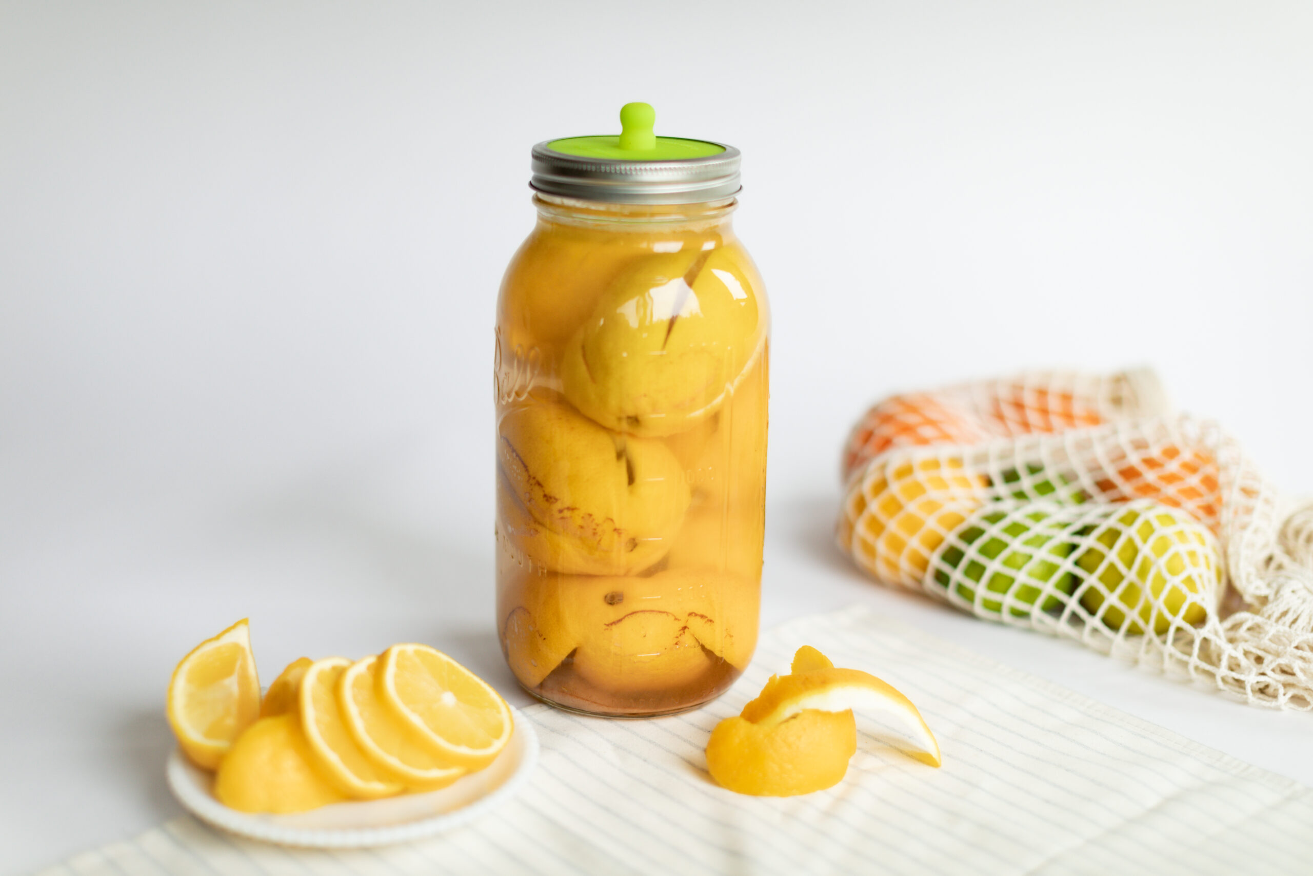 Jar of fermented lemons on a counter with a bag of citrus and sliced lemons on a plate