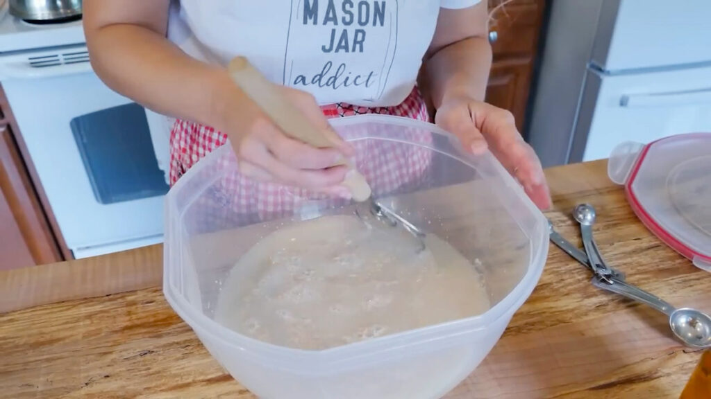 Hands using a dough whisk to mix yeast and water together in a large plastic bowl.
