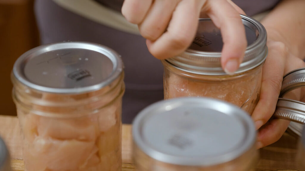 A woman's hands adding canning lids to jars of raw packed chicken.