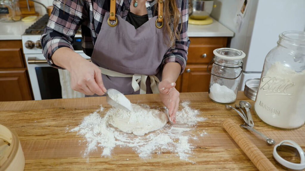 A woman sprinkling flour over a ball of bread dough.