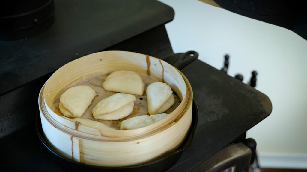 A basket of cooked bao buns in a wooden basket on a wood stove.