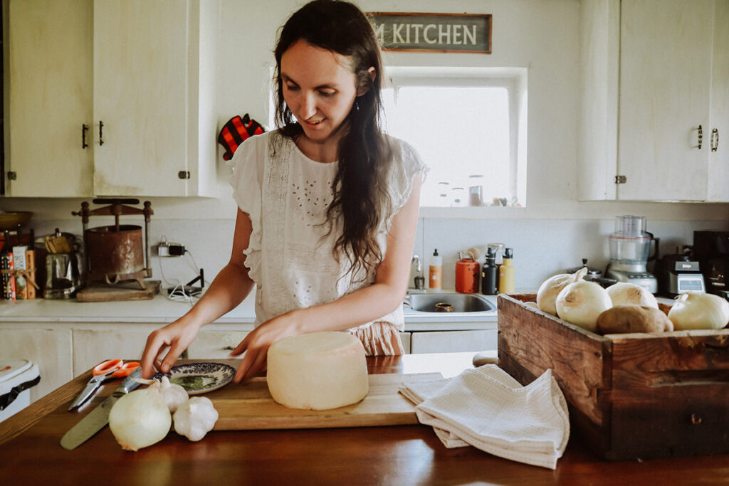 A woman in the kitchen with a wheel of homemade cheese on a cutting board.