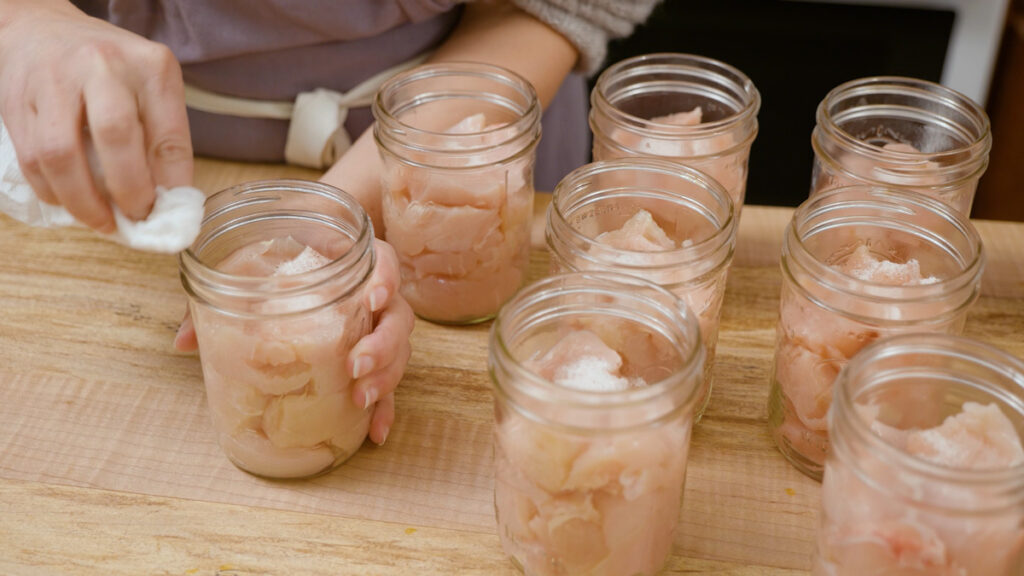A woman's hands wiping the rims of jars filled with raw chicken.