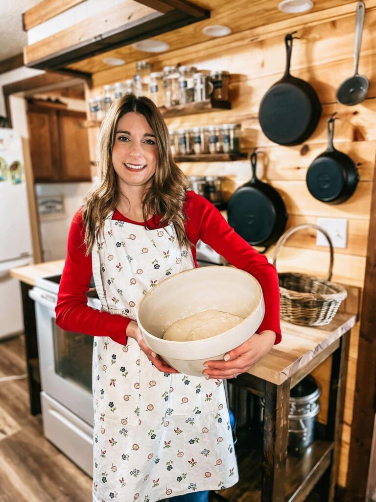 A woman in a kitchen holding a large bowl filled with bread dough.
