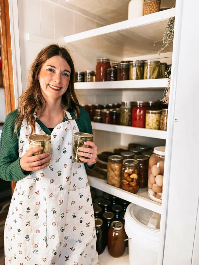 A woman holding two jars standing next to a pantry lined with home canned food.