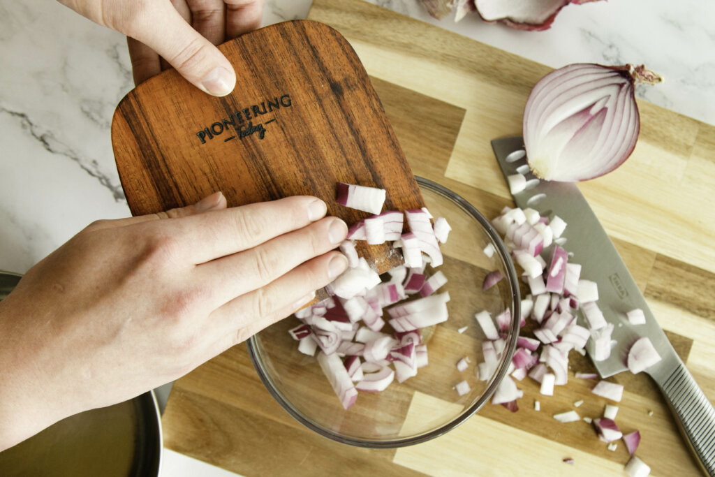 Wooden bench knife scooping onions up and putting them into a bowl.