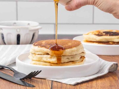 Three pancakes stacked on a white plate with maple syrup drizzling over the top.