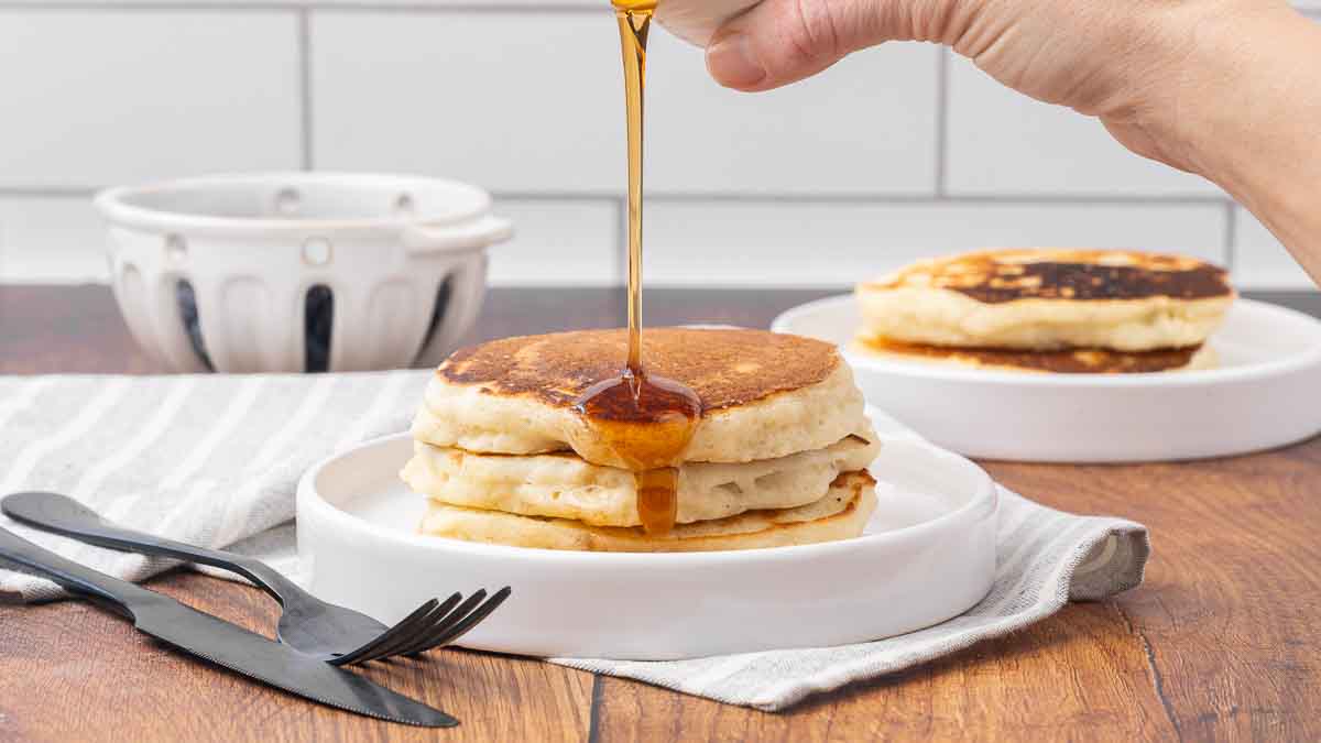 Three pancakes stacked on a white plate with maple syrup drizzling over the top.