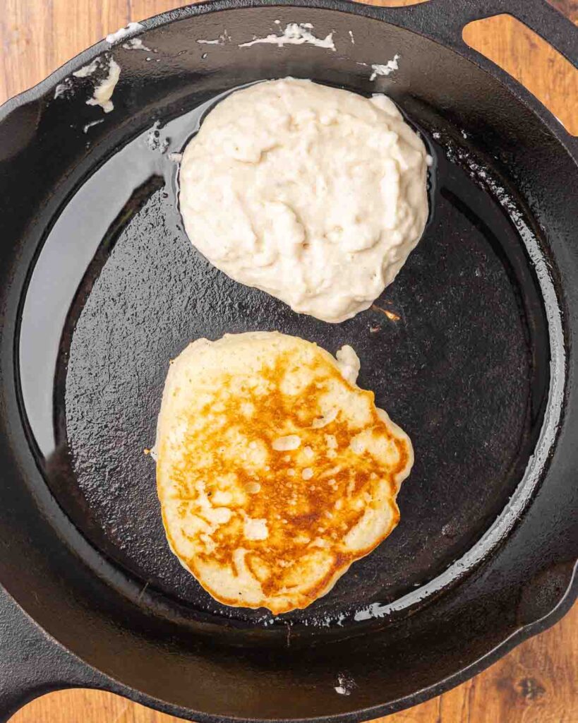 Two pancakes cooking in a cast iron skillet.
