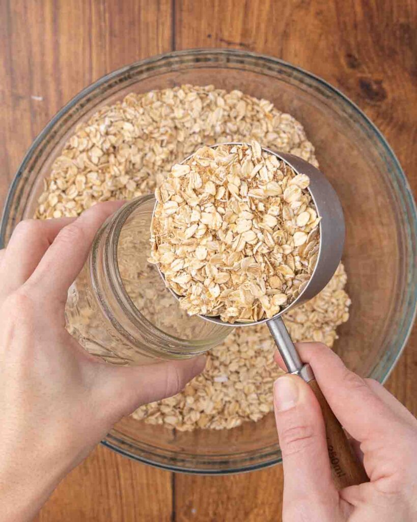 A measuring cup scooping instant oatmeal mix from a glass bowl into a glass jar.