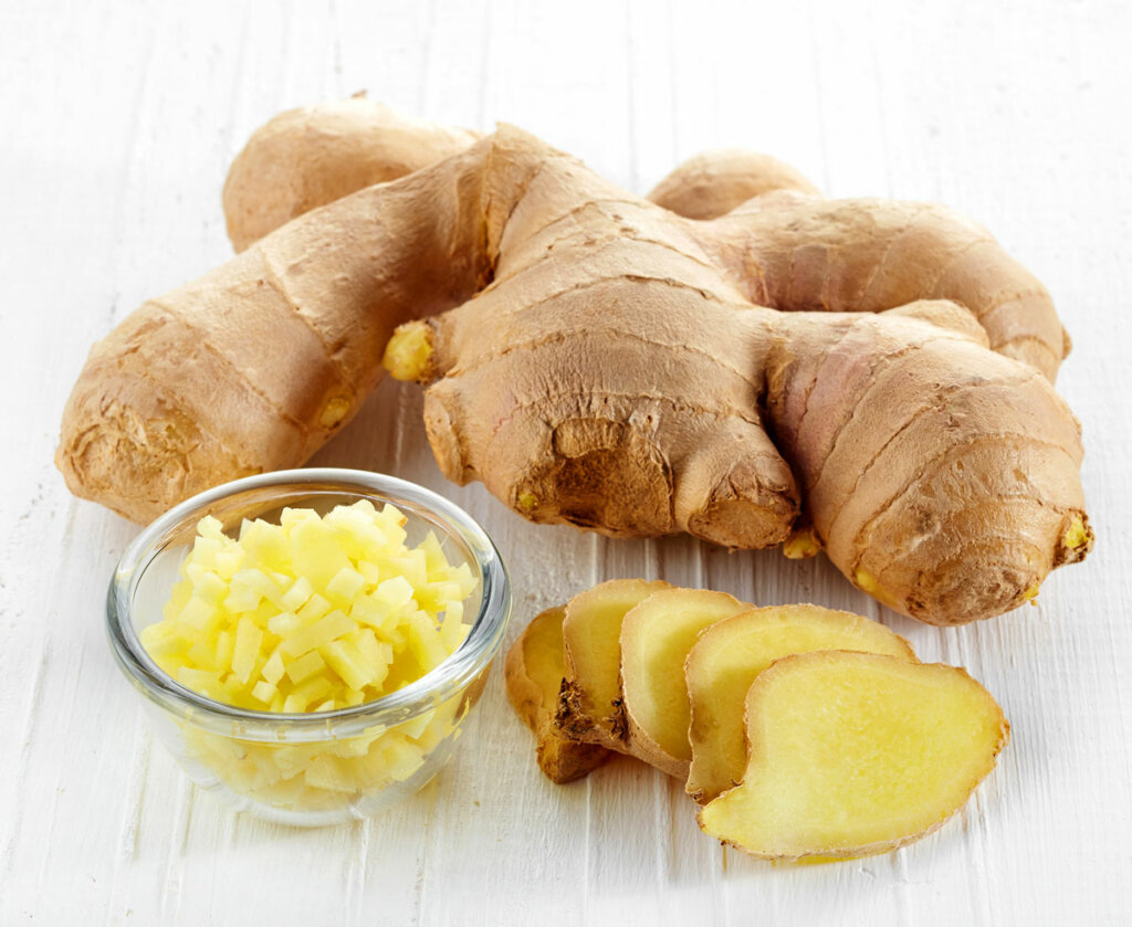 Ginger root on a white table top, sliced ginger root next to it and minced ginger root in a glass bowl.