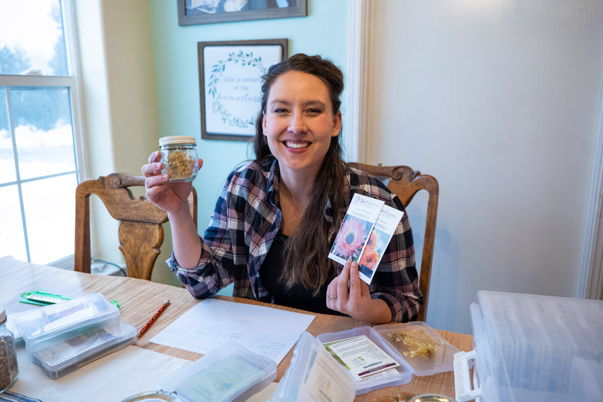 A woman sitting at a table with jars of dried herbs and flower seed packets.