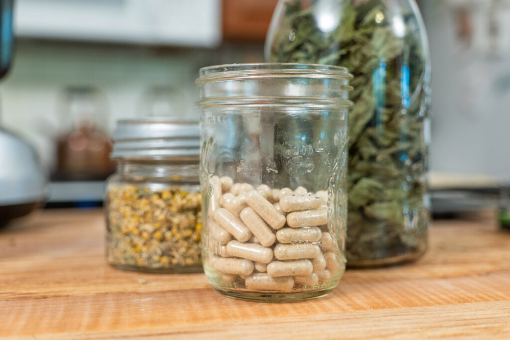 A jar of herbal capsules with jars of dried herbs on a wooden counter.