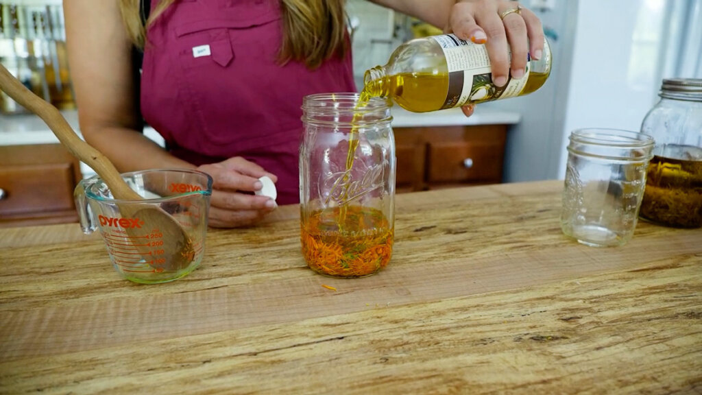 A woman pouring olive oil into a jar of dried calendula flowers.
