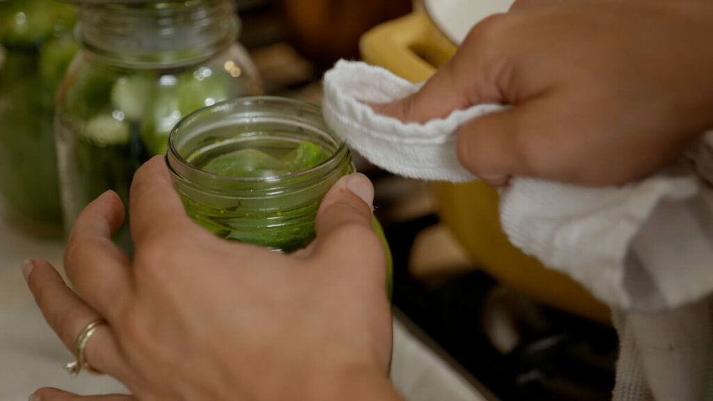 A towel wiping the rim of a canning jar filled with pickles.