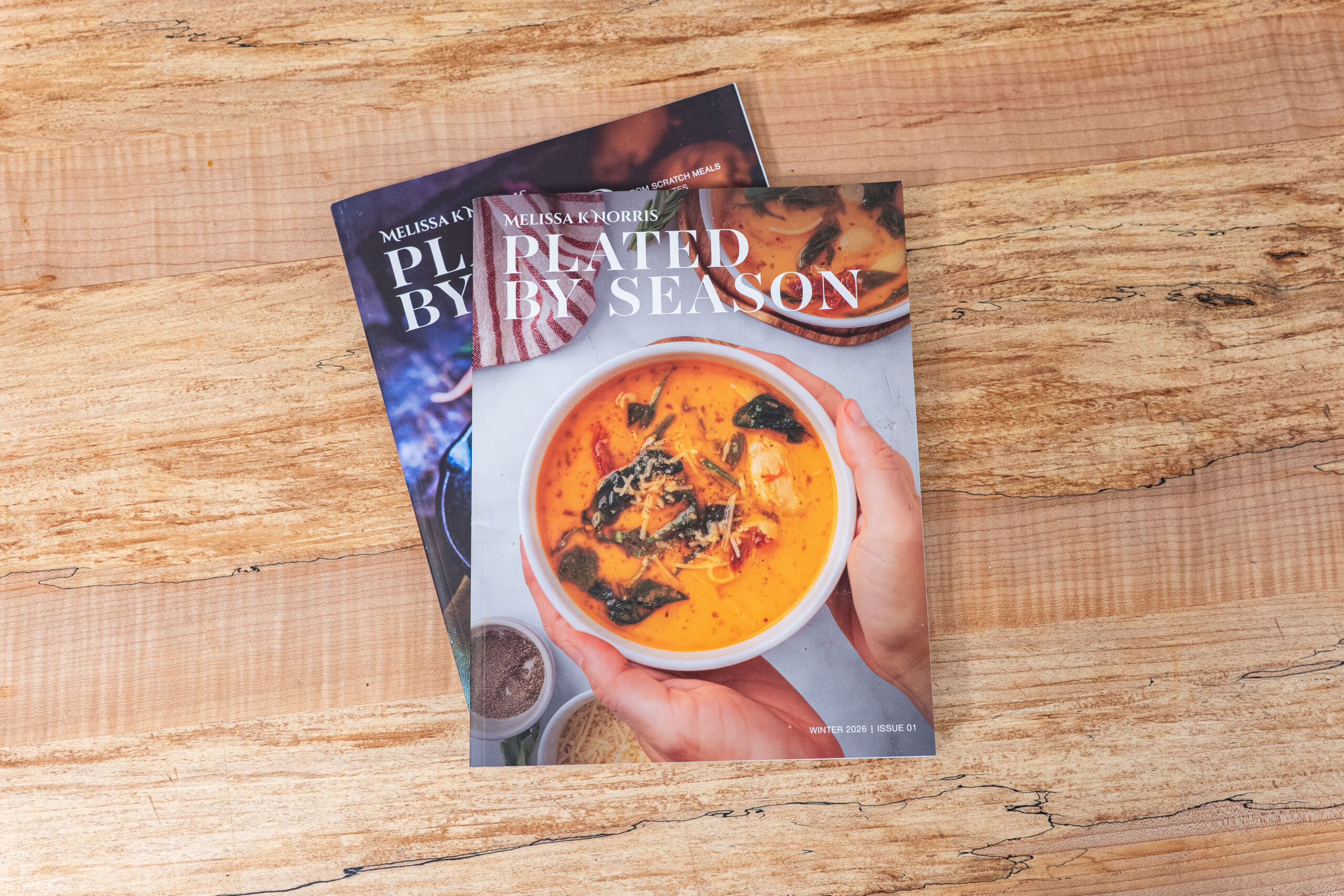Two issues of Plated by Season Magazine stacked on top of one another on a butcher block countertop