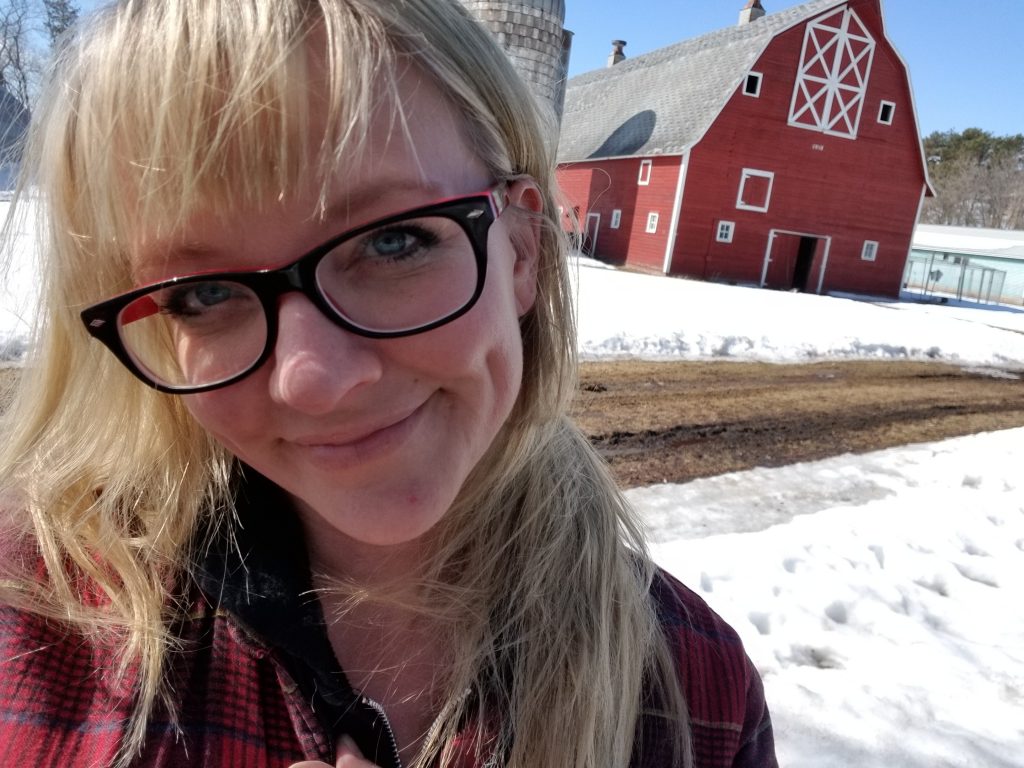 A woman standing in front of a big red barn, with snow on the ground.
