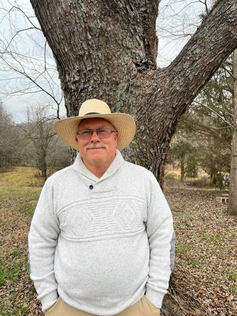 A man in a white shirt and hat standing by a large nut tree.