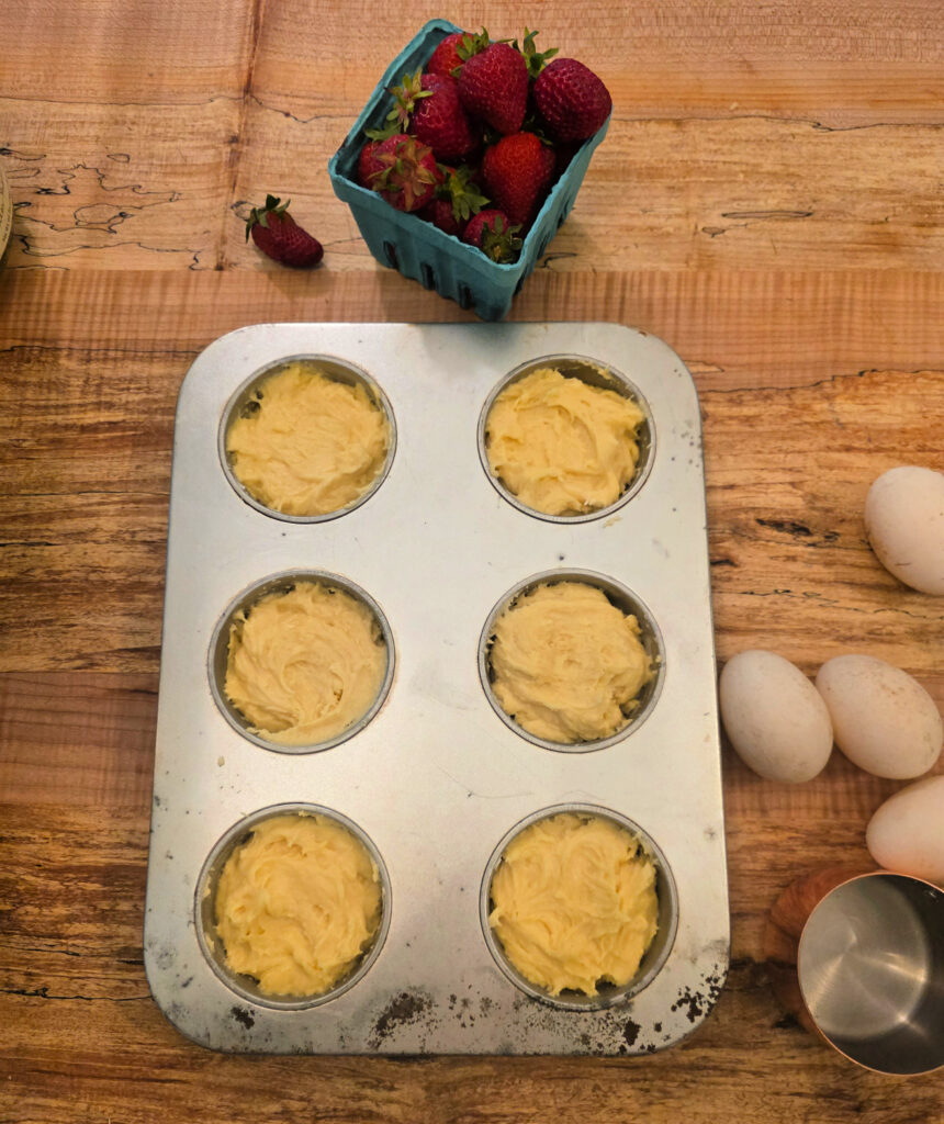 jumbo muffin tin filled with shortcake batter on counter top with strawberries and eggs