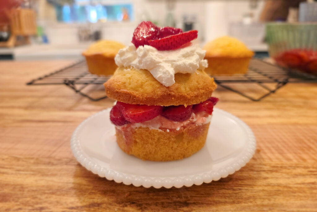 A homemade strawberry short cake on a white plate sitting on a wooden counterop with short cakes cooling on a wire rack in the background