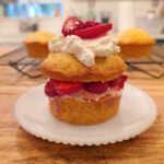 A homemade strawberry short cake on a white plate sitting on a wooden counterop with short cakes cooling on a wire rack in the background