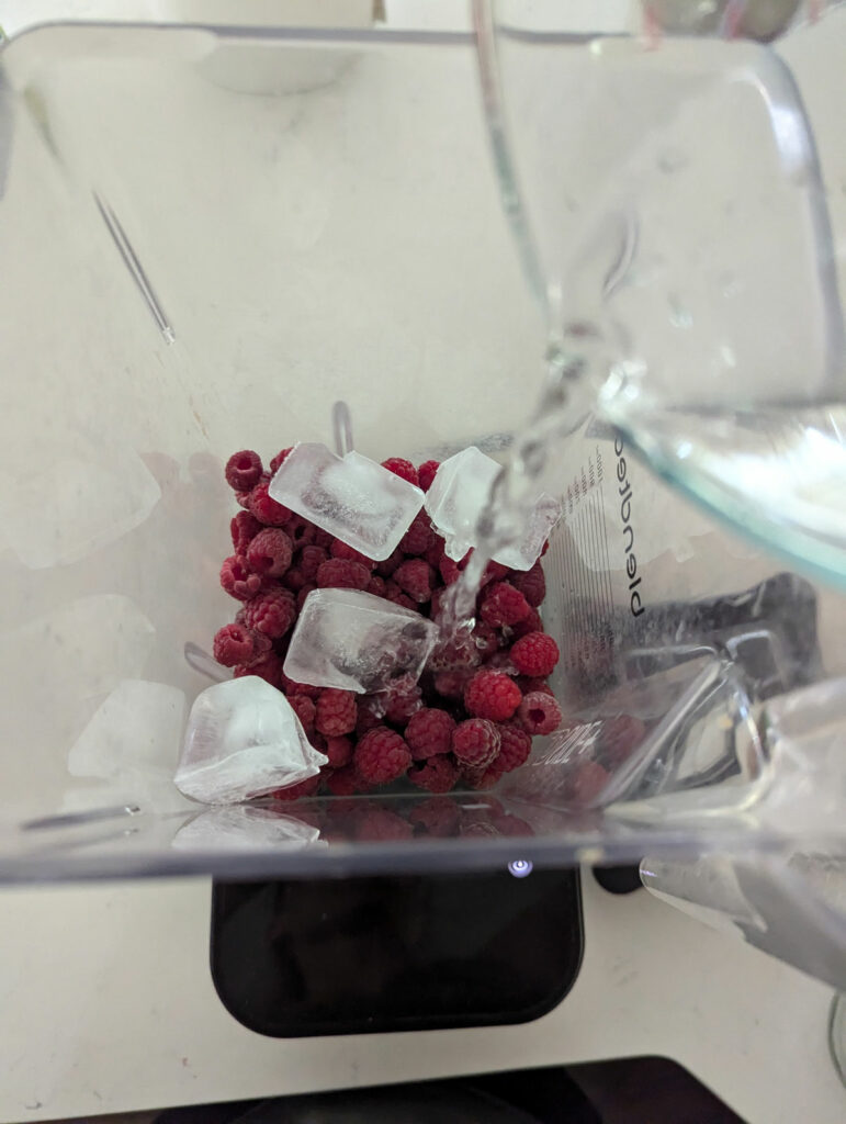 Raspberries and ice cubes in a blender with water being added.