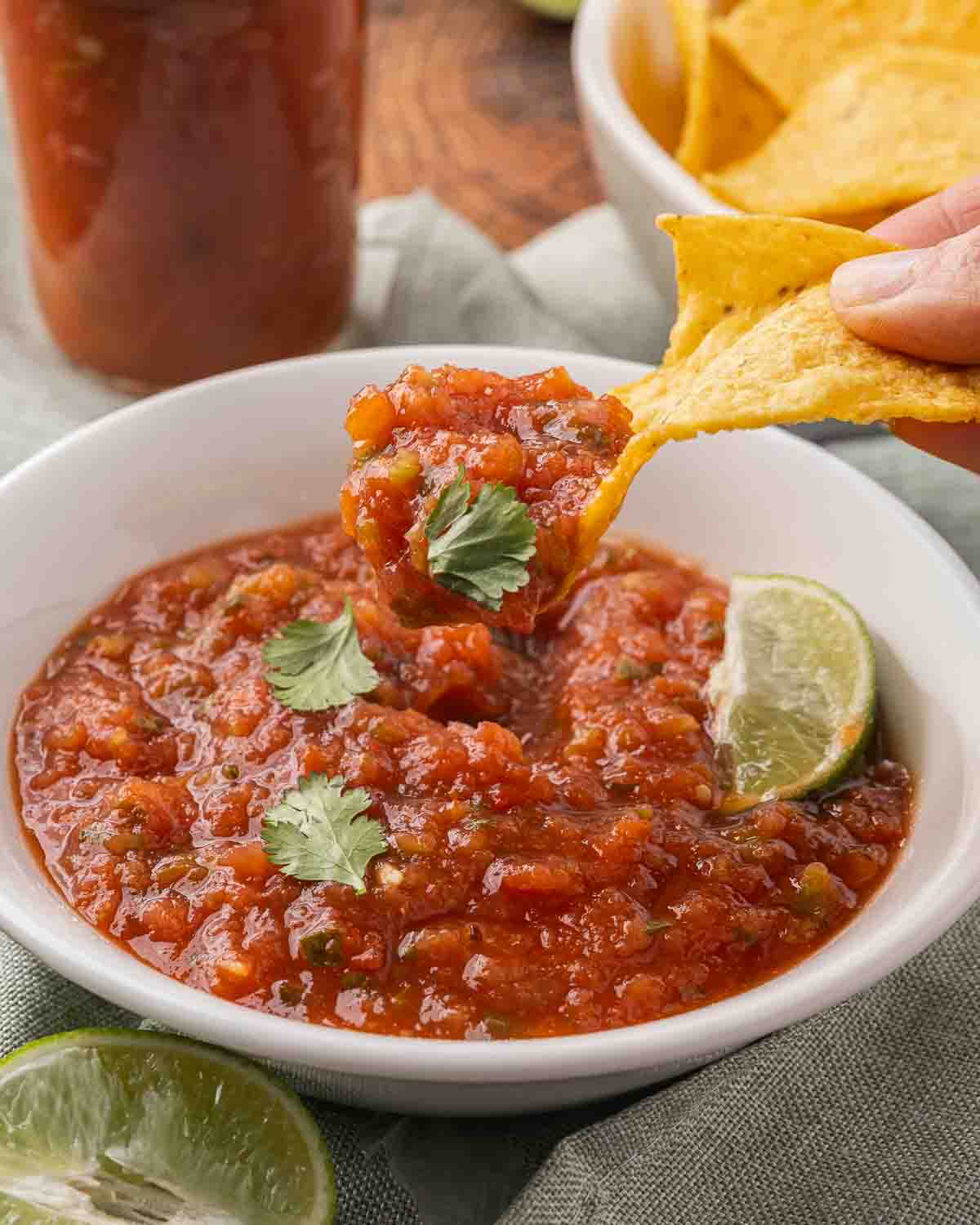 A white bowl full of fermented salsa garnished with cilantro, a wedge of lime and a tortilla chip dipping a bite.