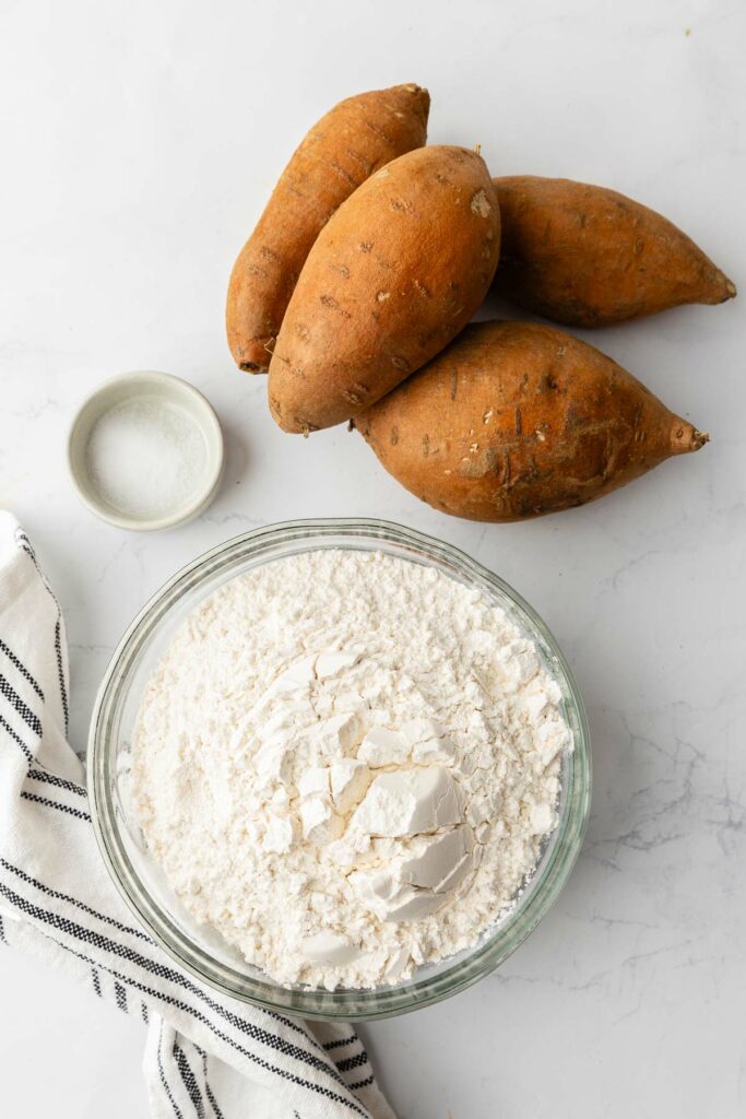 A bowl of flour, a small bowl of salt and sweet potatoes sitting on the counter.