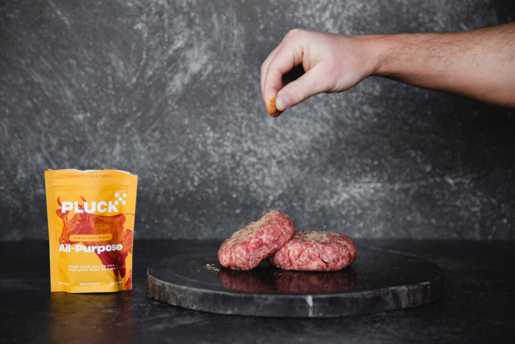 A hand seasoning raw hamburger patties with Pluck Seasoning.