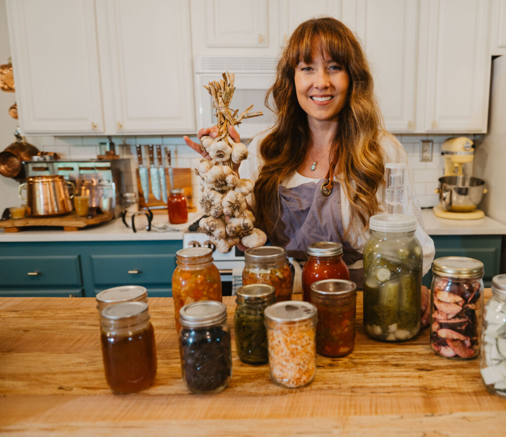 A woman with jars of preserved food and braided garlic in the kitchen.