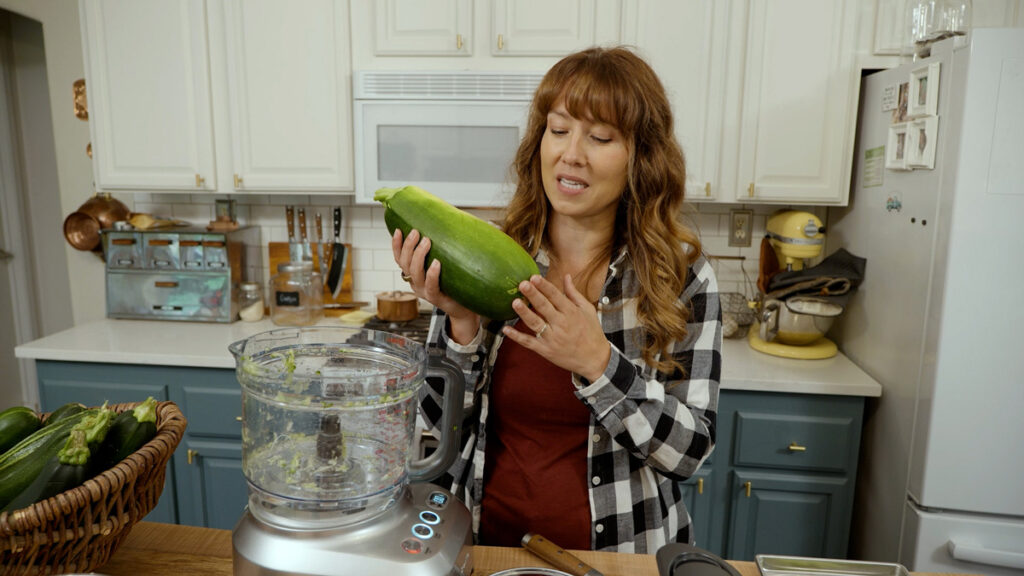 A woman in the kitchen holding up a large zucchini.