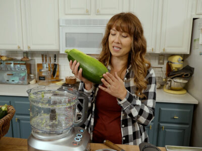 A woman in the kitchen holding up a large zucchini.