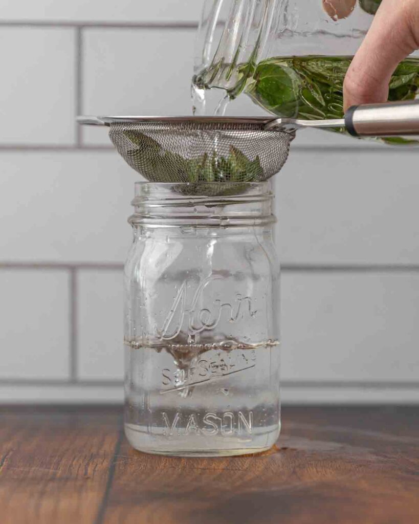 A hand pouring a jar full of mint and vodka through a strainer.