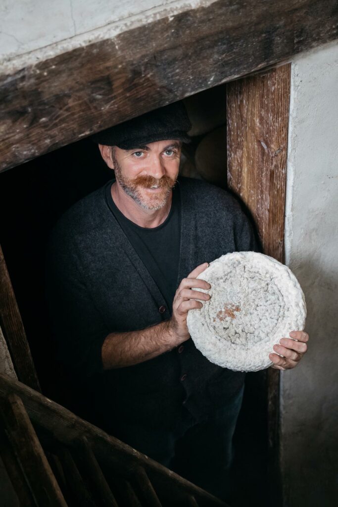 A man coming out of a cellar or cheese cave with a wheel of cheese.