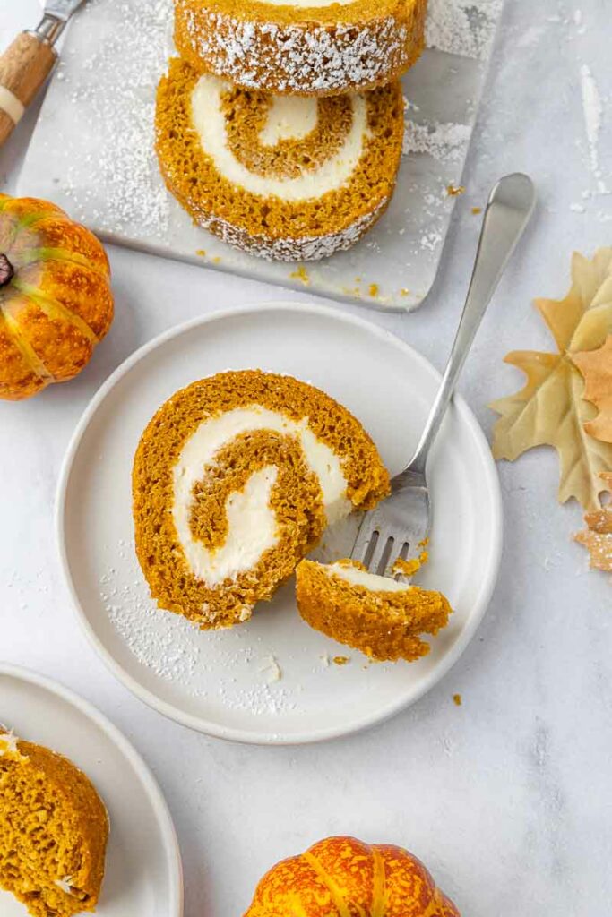 A slice of pumpkin roll on a white plate with a fork taking a bite.